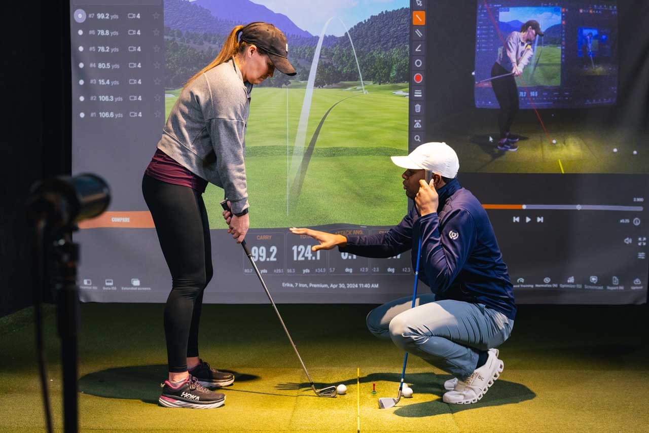 A Five Iron Golf coach guiding a member's swing on the simulator, with live shot data on screen behind them